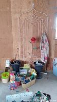 Photo shows a white wire trellis propped against a wall with various garden items in front including baskets, planters, tools, and books.