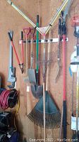 Assorted hand tools hung on wall: large metal tine rake, plastic fan rake, pitch fork, shovels and pipe wrench