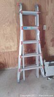 Aluminum multi-position ladder with red locks, folded to a smaller position leaning against wooden wall.