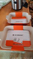 Two new rectangular Le Creuset stoneware cooking dishes stacked, showing brand labels and handles with ridges.