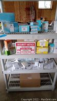 Shelf displaying various boxed security system components including cameras, transceivers, PC interfaces, and lamp modules along with a printed security sign.