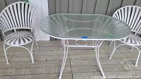 View of white metal framed table with square textured glass top and two matching white metal chairs with curved slatted backs and seats on wooden deck