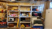 Three utility shelves in basement setting filled with various household items like fabric, sewing machine, bins, and tools.