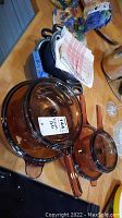 Photo showing two amber Vision glass pots with lids and handles placed on a wooden surface, along with other kitchen items in background.