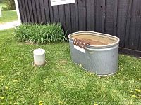 Photo of galvanized steel oval water trough with some wear and rust, outdoor setting on grass beside black wall