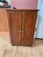 Front view of the wooden cabinet showing two paneled doors with silver knobs on a hardwood floor.