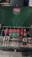 Green Coleman two-burner propane camping stove with red fuel tank and metal grate.