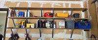 Three wall-mounted shelves in a garage with bottles of engine oils, linseed oil, Coleman fuel can, yellow plastic tote, black small toolbox, red gas can, and other small containers and boxed items spread across the shelves.