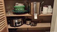 Shelves with kitchen appliances and cookware including lidded metal stock pot, Crockpot slow cooker, ceramic dishes, small pot and cookie press