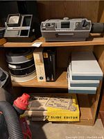 Shelf displaying two slide projectors along with multiple slide storage containers and boxed slide editor.