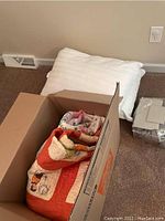 Quilts and pillows partially packed in box, showing colorful patterns and white pillows nearby.