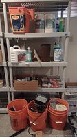 Wide view of shelving unit with cleaning and household items including Borax, paper towels, spray bottles, and containers, with three orange buckets on the floor below containing various tools and items.