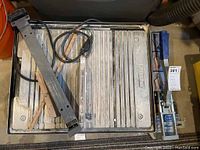 Top view of Plasplugs tile saw showing ridged cutting surface, electric cord, metal arm, and blue handle.