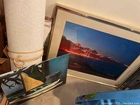 Stacked artworks: large metal-framed photograph behind small boat interior painting