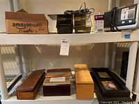 Shelf with multiple wooden and black boxes, picture frames, and an Amazon box labeled 'Graphics'