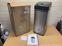 Photo of new 13-gallon stainless steel touchless trash can next to its original box and instruction manual on a wooden table.