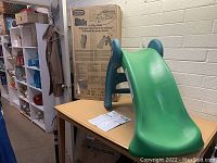 Green plastic Little Tikes toddler slide with original box and instruction manual on table in storage room.