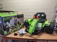 Huffy Electric Green Machine 360 kids' electric kart shown with original packaging box, charger, and manual on a wooden table against a brick wall.