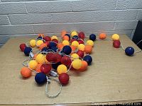 Large string of colorful ball lights in red, orange, yellow, and blue colors spread on a table against a grey brick wall.