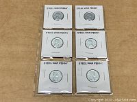 Six 1943 Steel War Pennies encased in individual white cardboard holders labeled 'STEEL WAR PENNY' arranged in two columns of three coins each, placed on a flat surface.