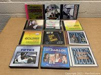 Photo showing 11 assorted music CDs on a table, featuring various genres like disco, oldies, blues, and 50s music.