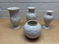 Group photo of four Lenox porcelain vases showing front view with relief floral designs and gold accents on a brown surface against a white brick background.