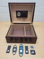 Open wooden cigar humidor box showing interior with thermometer and hygrometer on lid, with five metal cigar cutters and one round cigar punch laid out in front.