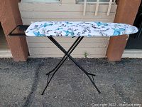 Front side view of ironing board showing hummingbird-patterned fabric cover and iron rest on black metal frame.