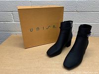Pair of black women's ankle boots with silver chain detail displayed with Unisa branded yellow box.