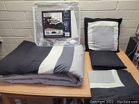 Comforter folded on table, two pillow cases, fitted sheet, flat sheet, pillow with geometric color block design boxed in plastic packaging in background.