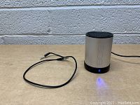 Silver and black 808 Bluetooth speaker powered on with blue indicator light showing, placed next to black 3.5mm auxiliary audio cable on wooden surface against brick wall.