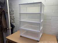 Clear four-drawer plastic storage unit with wheels on a wooden table against a white cinder block wall.