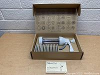 Cookie press kit in box showing cookie press gun, 11 design discs, and instruction booklet.