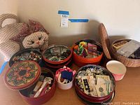 Overview image of fabric cat pincushion, various decorated tins filled with sewing supplies including buttons, threads, ribbons, and a wicker basket with packaged notions.