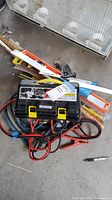 Overview photo showing black hard plastic case with yellow clasps, jumper cables with red and black clamps, several measuring rulers, and other tools on concrete floor.