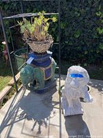 Elephant and lion sculptures positioned outdoors with bicycle rack in background. Elephant holds succulent plant.