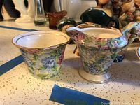 Photo of floral patterned porcelain creamer and sugar bowl with detailed blue, green, and purple flower designs on counter with other figurines in background.