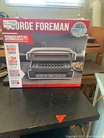Front view of the George Foreman Smokeless Grill box showing grill image, brand name, and key features including smokeless grilling and faster cooking.