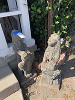 Photo of two weathered concrete lion sculptures standing on ground near a white fence and garden vegetation.