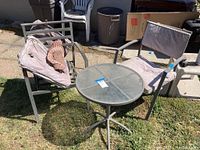 Outdoor shot showing round side table with glass top and two metal frame patio chairs on grass. Weathered condition visible.
