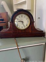 Front view of the brown wooden mantel clock showing the clock face with black numbers and hands, brass bezel, and electric cord.