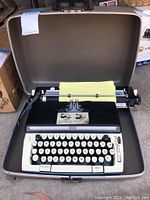 Open typewriter showing keyboard keys, paper roller with yellow lined paper, and control dials within the original case.