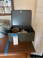 Gray metal box on wooden surface with lid open showing some of the contents and a MaxSold tag.