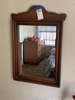 Front view of the dark wood framed rectangular mirror with sculpted top edge hanging on wall reflecting bedroom furniture.
