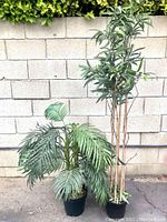 Six bamboo stalks with attached green leaves arranged in a black pot, shown against a concrete block wall, with a smaller artificial palm plant visible next to it.