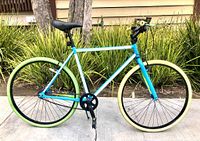 Left side view of the full KENT single speed fixed gear bike showing the blue glow in the dark frame, black seat, green rear tire, cream front tire, and yellow handlebar grips.