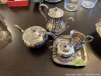 Five piece silver tea set displayed on a table including teapot, sugar bowl with lid, creamer, and tray