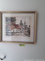 Framed watercolor painting of a village scene showing cottages with tile roofs and a church steeple, hung on a wall.