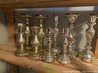 Nine assorted metal candle sticks displayed on a wooden shelf under a table, showing different sizes, designs, and finishes.