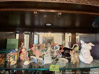 Photo of assorted porcelain and ceramic figurines arranged on a glass shelf under wood cabinet. Includes animal figurines, female figurine, decorative containers.